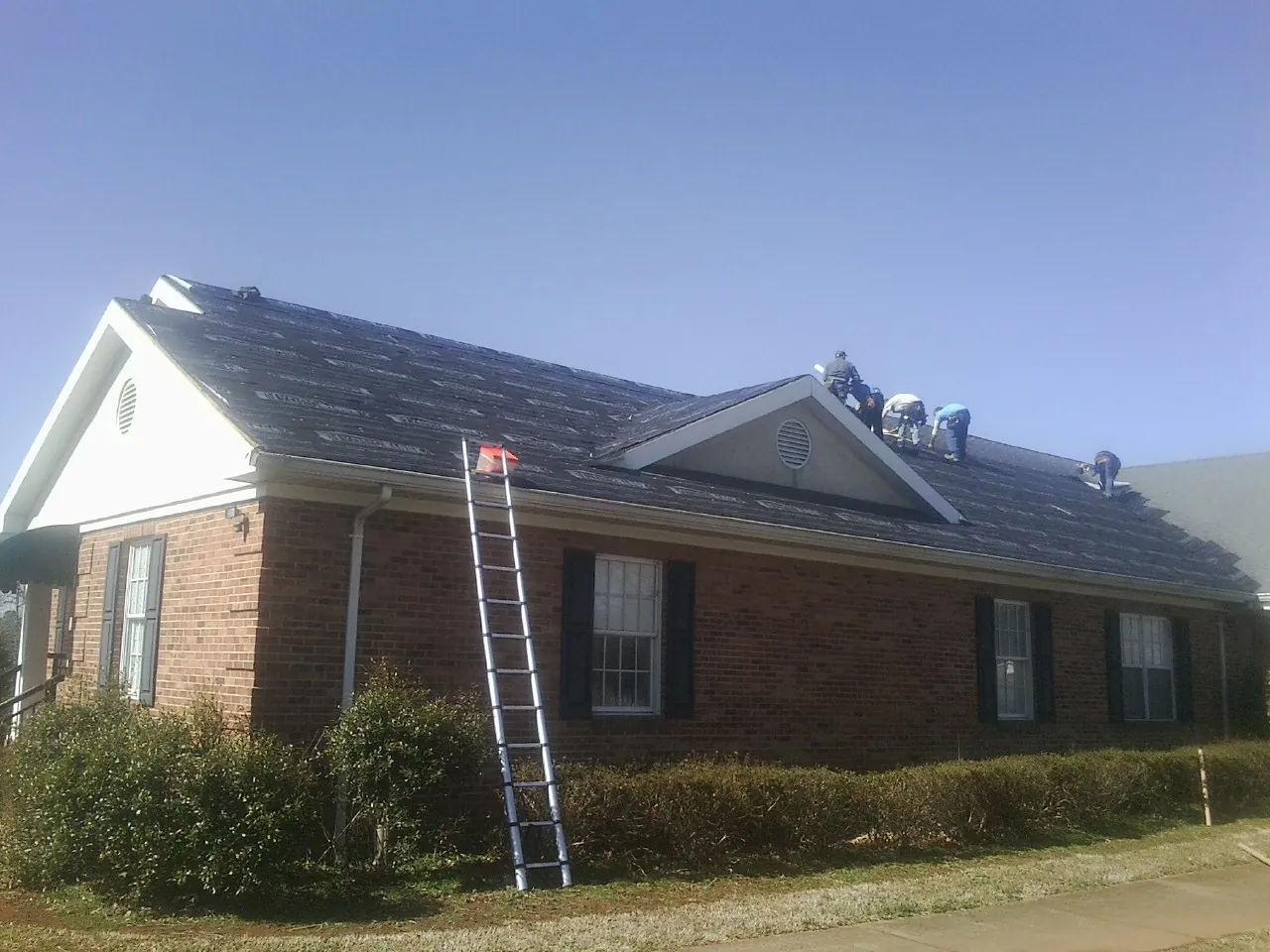 Crew tearing shingles off a brick ranch roof with a ladder set — Emergency Roof Repair work by our crew in Fort Lupton