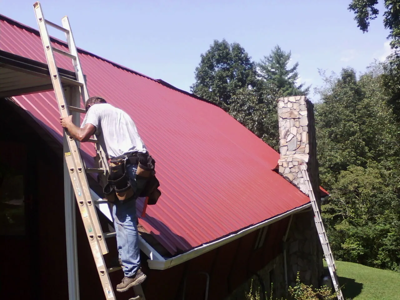 Roofer on a ladder beside a red metal roof and stone chimney — Soffit & Fascia Repair by Roofing Brothers in Fort Lupton